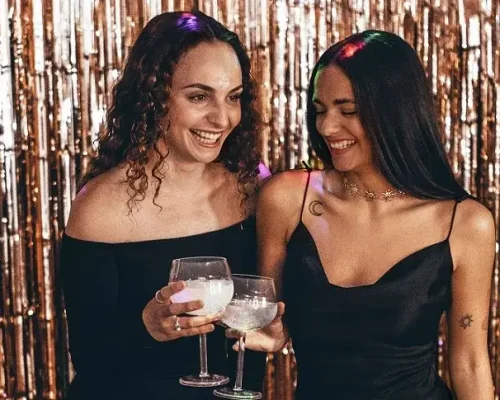 Dos mujeres vestidas de negro, con una copa en la mano, durante una fiesta. Posan frente a un fotomatón | alquiler de fotomatones